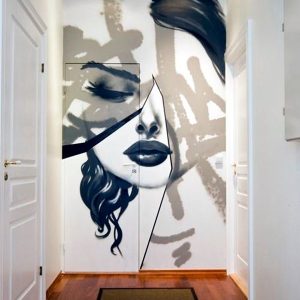A striking black-and-white mural of a woman's face with abstract geometric elements painted on a hallway wall. The artwork features bold brush strokes, fragmented lines, and a mix of realism and abstraction, creating a dramatic and modern artistic statement. The face appears divided yet seamlessly connected, blending surrealism with contemporary street art aesthetics.