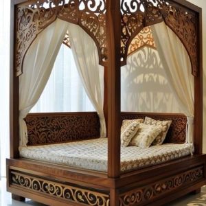 Intricately carved wooden canopy bed with sheer white drapes and traditional patterned bedding, placed in a modern, sunlit room.