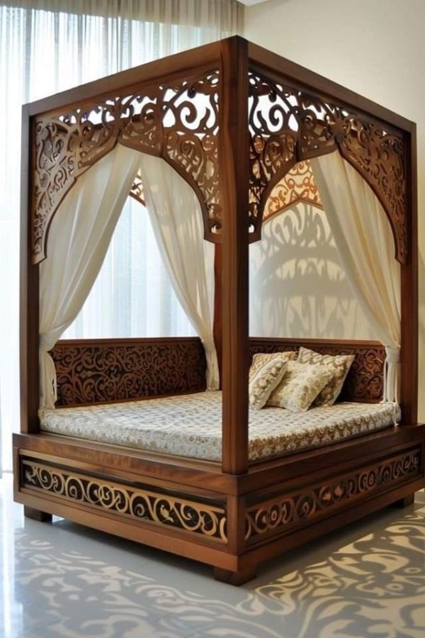 Intricately carved wooden canopy bed with sheer white drapes and traditional patterned bedding, placed in a modern, sunlit room.